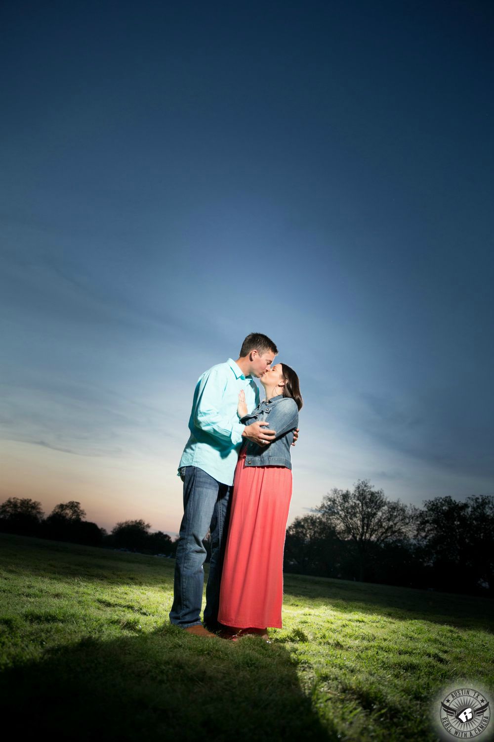 Brunette tan girl wearing a long pink dress in a denim jacket kisses a taller guy with dark hair and a light blue long sleeve button up shirt and blue jeans standing in a green grassy field at dusk with a big blue and orange Texas sky in the background of this enchanting engagement photo at Zilker Park in Austin. 
